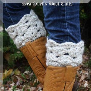 May include: A pair of light grey crochet boot cuffs with a sea shell pattern, worn over brown leather cowboy boots. The boots are on fallen leaves. Text reads "Sea Shells Boot Cuffs" and "Designs by Mamta".