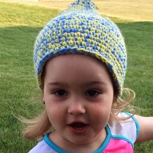 May include: A handmade crochet hat in blue and yellow, with a pointed top. The hat is worn by a young child. The child is wearing a pink and white shirt with blue trim. The text "Designs by Mamta" is visible at the bottom of the image.