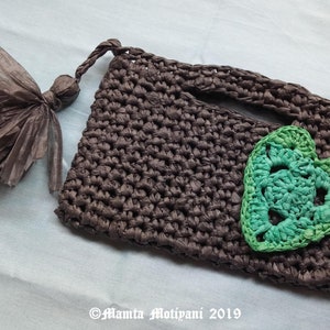 May include: A handmade brown crochet clutch with a green heart applique. The clutch has a matching tassel and a handle. The design is simple and elegant, perfect for carrying essentials. The clutch is made of a durable material.