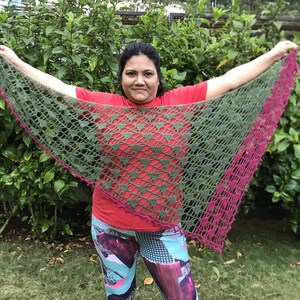 May include: A handmade, olive green crochet shawl with a triangular shape. The shawl features a delicate openwork design and a vibrant fuchsia border. The shawl is held up against a background of green foliage.