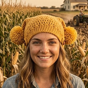 Peut inclure: Une femme souriante porte un chapeau jaune crocheté avec des pompons, une salopette en jean et une chemise en jean dans un champ de maïs. Un tracteur laboure un champ en arrière-plan, avec une maison visible au loin.