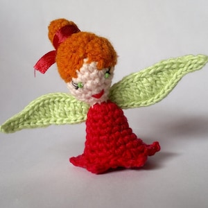 May include: A small crocheted fairy doll featuring a red dress, light green wings, and orange yarn hair styled in a bun with a red ribbon. The doll has a pale face with green bead eyes and a stitched red mouth, crafted with textured yarn.