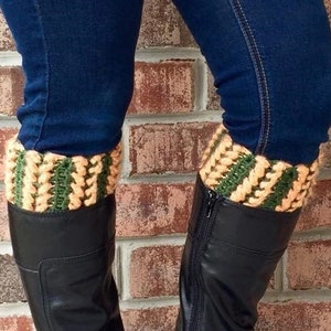 Op de afbeelding: Zwarte kniehoge laarzen met gespen en ritsen, gecombineerd met een blauwe jeans. De laarzen worden geaccentueerd door gebreide beenwarmers met groene en beige strepen. De achtergrond is een bakstenen muur.