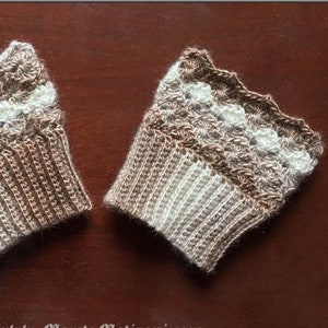Op de afbeelding: Twee handgemaakte gehaakte polswarmers in bruin en wit. De polswarmers hebben een geschulpte rand en een ribgebreid ontwerp aan de onderkant. Ze worden gepresenteerd op een bruin oppervlak.