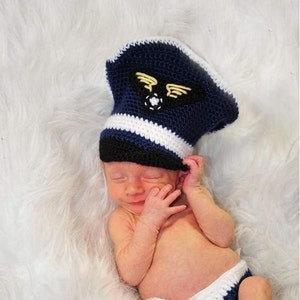 May include: A navy blue and white crocheted pilot hat with a black brim and a yellow embroidered wing emblem, paired with matching navy blue and white crocheted diaper cover shorts. The hat features a distinct pilot cap shape with a white band.