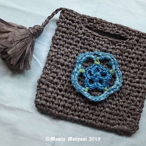 May include: A handmade brown woven bag with a blue and green floral applique. The bag has a matching tassel and a small handle. The bag is made of a woven material and has a square shape.