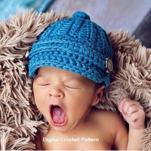 Könnte beinhalten: Ein Baby mit einer leuchtend blauen Häkelmütze mit Knopfdetails. Das Baby gähnt. Die Mütze liegt auf einem weichen, flauschigen, braunen Untergrund. Der Text "Digital Crochet Pattern" befindet sich am unteren Bildrand.