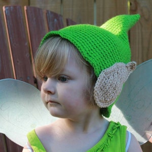 May include: A child wearing a green crocheted elf hat with tan ear details and clear fairy wings. The hat has a pointed top and a brim that covers the forehead. The wings are attached to the back of the child's outfit.