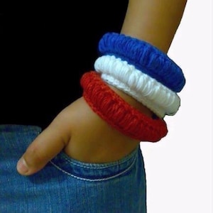 May include: Three crochet bracelets in red, white, and blue are stacked on a wrist. The bracelets are thick and appear handmade. The person is wearing blue jeans and a black shirt. The text "Designs by Mamta 2010" is visible.