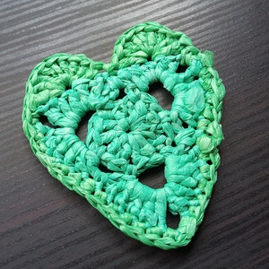 May include: A handmade, heart-shaped coaster in shades of green. The coaster is crocheted with a textured, woven appearance. The design features openwork circles within the heart shape, adding visual interest. The coaster is placed on a dark wood surface.