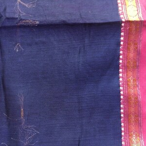May include: A dark blue fabric with a pink border and gold accents. The fabric appears to be a saree or similar garment, with visible texture and some loose threads. The pink border has a gold and white design.
