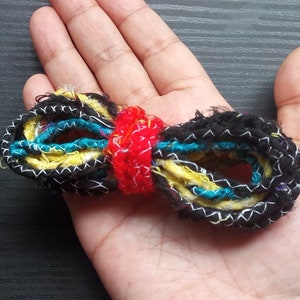 May include: A bow-shaped accessory crafted from multiple strands of textured yarn in black, yellow, and teal, featuring a bright red center band. The black outer strands are detailed with a white zigzag stitch pattern throughout the entire length.
