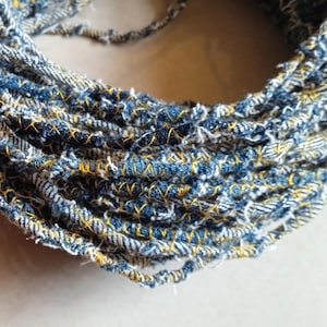 May include: A close-up shot of a coiled textile item, likely handmade. It's constructed from strips of blue denim with yellow stitching, forming a textured, circular shape. Frayed edges give a rustic, handcrafted appearance. It could be a decorative item or craft material.