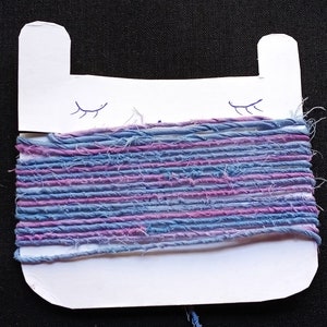 May include: A white cardboard bobbin shaped like a cat head with two ears and drawn closed eyes, wrapped with multiple strands of textured yarn in alternating shades of light blue, periwinkle, and purple, featuring a frayed and rustic appearance.
