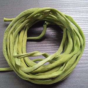 Op de afbeelding: Een opgerolde bundel olijfgroene stoffen stroken, waarschijnlijk lint of sierband, ligt op een donkere ondergrond. De stof lijkt een zacht materiaal te zijn, mogelijk katoen of een vergelijkbaar textiel. De stroken zijn netjes opgerold.