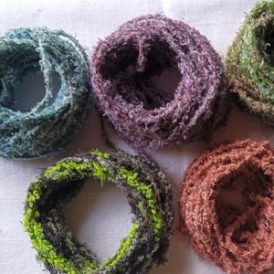 May include: Five circular skeins of textured yarn arranged in a group. The yarn colors include blue, purple, green, a mix of dark green and lime, and a solid copper-orange. Each skein features a fuzzy, frayed appearance with a loose, open-loop structure.