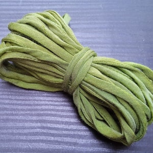 Op de afbeelding: Een bundel olijfgroene stoffen stroken, strak opgerold en vastgezet met een bijpassende stoffen band. Het materiaal lijkt zacht en licht getextureerd, wat suggereert dat het gebruikt kan worden voor knutselen of decoratieve doeleinden.