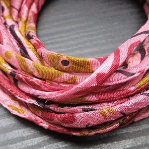 May include: A coiled fabric strip in shades of pink, featuring a floral design in red, yellow, and black. The material looks soft, possibly silk. The strip is arranged in a circular coil, likely for crafting or decoration.