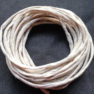 May include: Coiled bundle of off-white fabric strips with subtle gray stripes. The strips are tightly wound, creating a circular shape. The fabric appears to have a slightly textured surface. The background is a solid black.