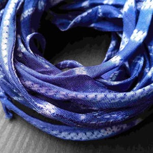 May include: A coiled length of blue fabric ribbon featuring a repeating white tie-dye pattern. The material has a smooth, slightly reflective texture and is wound into a circular shape, displaying multiple layers of the patterned textile strip.