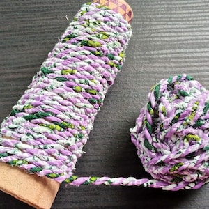Puede incluir: Un carrete de cinta de tela retorcida en tonos lavanda, blanco y verde, con una pequeña bola del mismo material al lado. La cinta está enrollada alrededor de un núcleo de cartón con una base marrón.