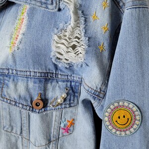 May include: Light blue denim jacket with distressed details, embroidered stars, a rainbow stripe, and a smiley face patch. The jacket has a frayed hem and a small bow pin.