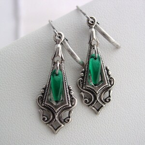 May include: A pair of silver-toned drop earrings with a vintage design. Each earring features a decorative frame with an emerald green, teardrop-shaped glass insert. The earrings have a hook closure.