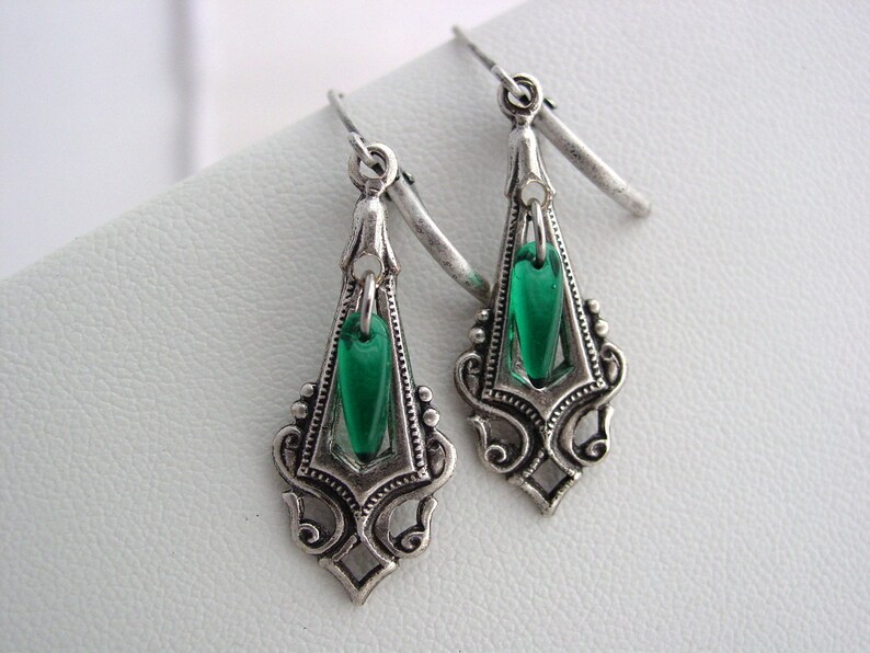 May include: A pair of silver-toned drop earrings with a vintage design. Each earring features a decorative frame with an emerald green, teardrop-shaped glass insert. The earrings have a hook closure.