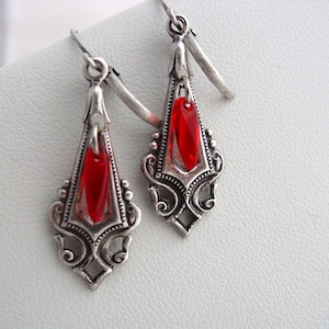 May include: A pair of silver dangle earrings with red teardrop-shaped glass beads. The earrings have a delicate, ornate design.