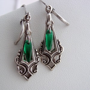 May include: A pair of silver-toned Art Deco style earrings. Each earring features an ornate, openwork design with a teardrop-shaped, emerald green glass bead suspended in the center. The earrings have a hook closure.