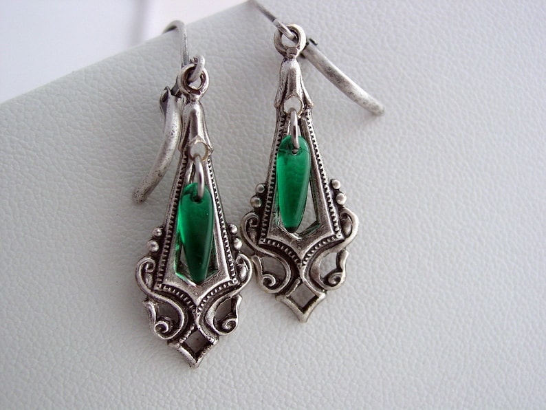 May include: A pair of silver-toned Art Deco style earrings. Each earring features an ornate, openwork design with a teardrop-shaped, emerald green glass bead suspended in the center. The earrings have a hook closure.