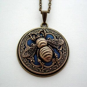 May include: A gold-toned pendant necklace features a round, blue enamel charm with a detailed, gold-toned bee design. The charm is surrounded by a decorative, gold-toned filigree border.