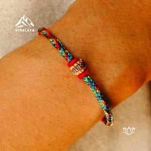 May include: A colorful braided bracelet with red, blue, green, and yellow threads. The bracelet features a central decorative element and red accents. The word "HIMALAYA" is printed on the upper left.