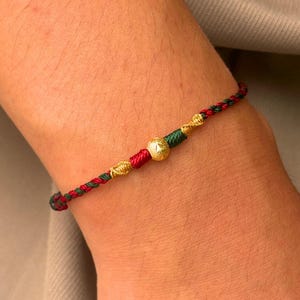 Pode incluir: Uma pulseira feita de cordão trançado vermelho e verde escuro, apresentando uma conta esférica dourada central com superfície texturizada, ladeada por dois pequenos nós decorativos em tom dourado de cada lado, presa no pulso.