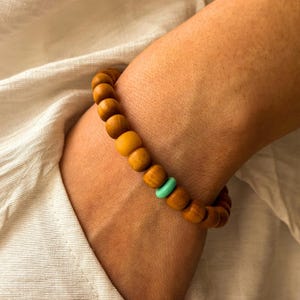 May include: Wooden bead bracelet with a light brown color and a single turquoise bead. The bracelet is made of round wooden beads and is worn on a wrist.