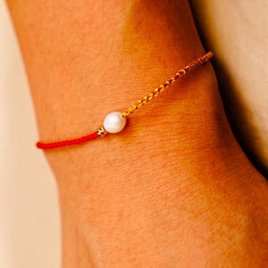 Può includere: Un braccialetto rosso e oro con una perla bianca. Il braccialetto è composto da un cordoncino rosso che si trasforma in una catena d'oro. La perla è al centro del braccialetto.