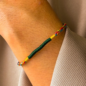 Può includere: Un braccialetto intrecciato con fili rossi, gialli, blu e verdi. Il braccialetto ha una sezione centrale verde scuro e dettagli dorati. Il braccialetto è indossato su un polso.