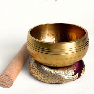 May include: A brass Tibetan mini singing bowl, with a wooden striker and cushion ring. The bowl has the text "Tibetan Mini Singing Bowl Handcrafted, Clear, Calming Resonance". The cushion is patterned.