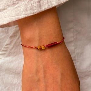 Könnte beinhalten: Ein rot-cremefarbenes Armband mit zwei gelben Perlen und einer roten rechteckigen Perle. Das Armband wird am Handgelenk getragen, mit einem weißen Hemd im Hintergrund.