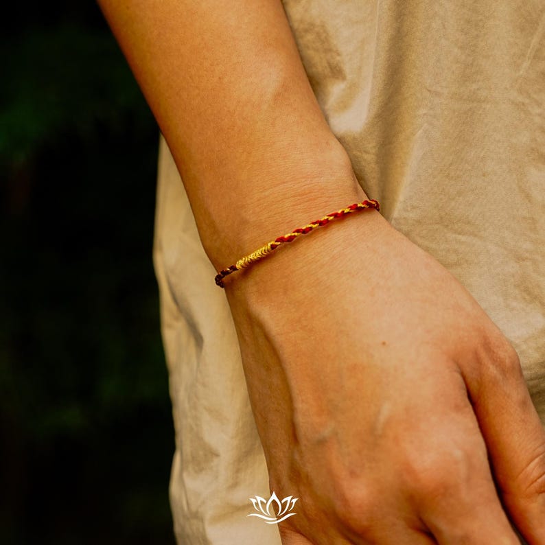 Puede incluir: Una pulsera trenzada con hilos rojos y amarillos. La pulsera se lleva en la mu&ntilde;eca, con la piel de color marr&oacute;n claro. El fondo est&aacute; borroso, con tonos verde oscuro y beige.