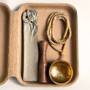 May include: A meditation set in a cardboard container. The set includes a gray stone incense holder, a wooden pestle, a beaded mala necklace, and a small brass singing bowl. The items are arranged in a brown, textured container.