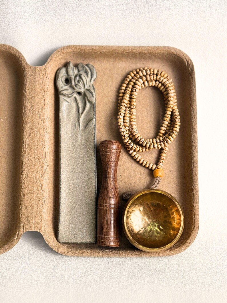 May include: A meditation set in a cardboard container. The set includes a gray stone incense holder, a wooden pestle, a beaded mala necklace, and a small brass singing bowl. The items are arranged in a brown, textured container.