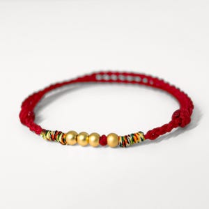May include: Red braided bracelet with four gold-colored beads and a single red bead. The bracelet features colorful woven accents near the ends. The bracelet is set against a white background.