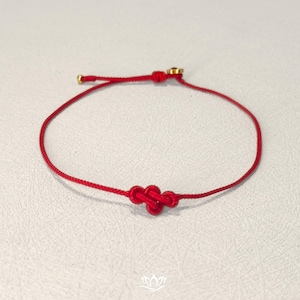 Pode incluir: Uma pulseira de fio vermelho com um n&oacute; decorativo no centro. A pulseira tem detalhes em metal dourado e &eacute; apresentada em uma superf&iacute;cie branca. Um acess&oacute;rio simples e elegante.