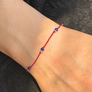 Può includere: Un braccialetto di filo rosso con tre piccole perline blu rotonde. Il braccialetto è indossato su un polso, con il filo rosso e le perline blu che contrastano con il tono della pelle. Il braccialetto è semplice e delicato.