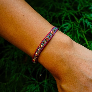 Puede incluir: Una pulsera colorida con base roja, adornada con detalles dorados y pequeñas gemas rojas redondas. La pulsera presenta un patrón turquesa y dorado en el centro, creando un accesorio vibrante.