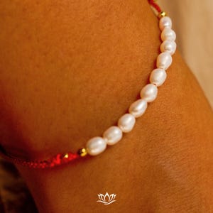 Peut inclure: Gros plan sur un bracelet compos&eacute; de perles blanches ovales. Les perles sont enfil&eacute;es sur un cordon rouge avec des d&eacute;tails dor&eacute;s. Le bracelet a un design simple et &eacute;l&eacute;gant, adapt&eacute; &agrave; un usage quotidien ou &agrave; des occasions sp&eacute;ciales.