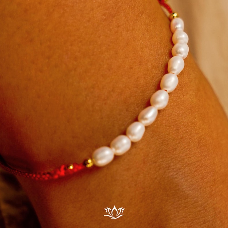 Peut inclure: Gros plan sur un bracelet compos&eacute; de perles blanches ovales. Les perles sont enfil&eacute;es sur un cordon rouge avec des d&eacute;tails dor&eacute;s. Le bracelet a un design simple et &eacute;l&eacute;gant, adapt&eacute; &agrave; un usage quotidien ou &agrave; des occasions sp&eacute;ciales.