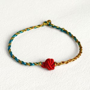 Può includere: Un braccialetto intrecciato con accenti oro, turchese e rossi. Il braccialetto presenta un design a nodo rosso centrale, con il cordoncino intrecciato che passa dall'oro al turchese. Il braccialetto è esposto su una superficie bianca.