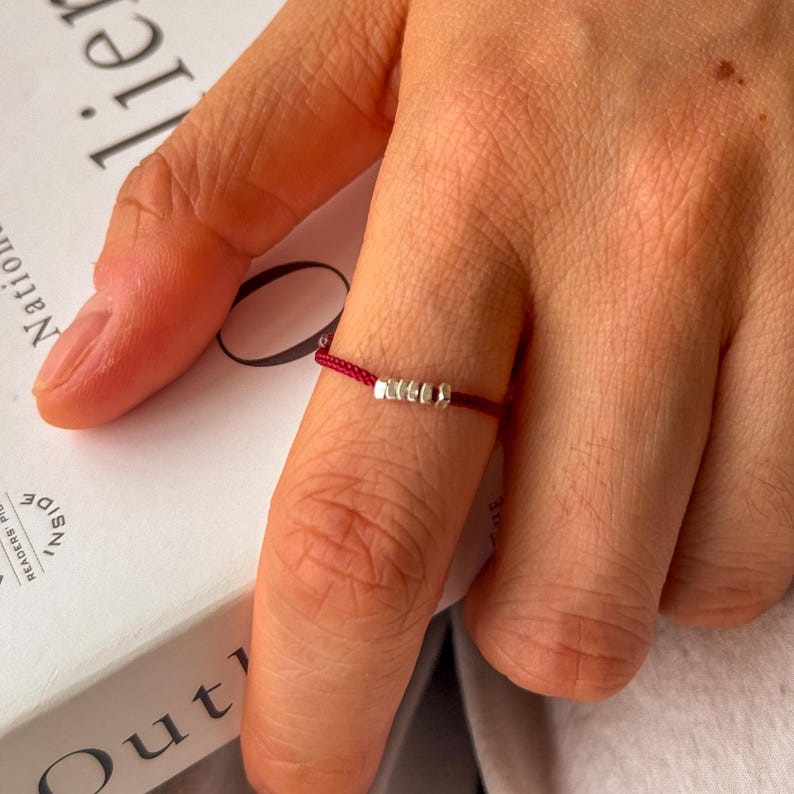 May include: A minimalist ring featuring a thin, deep red cord with three small, silver-colored beads. The ring is worn on a finger, showcasing its simple yet elegant design. The background includes a book and a person's hand.